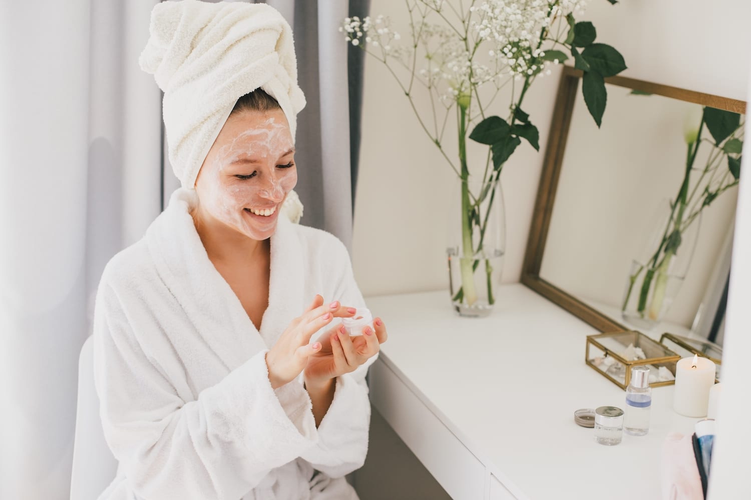 young woman in a bathrobe applying mosturizing skin cream to her face